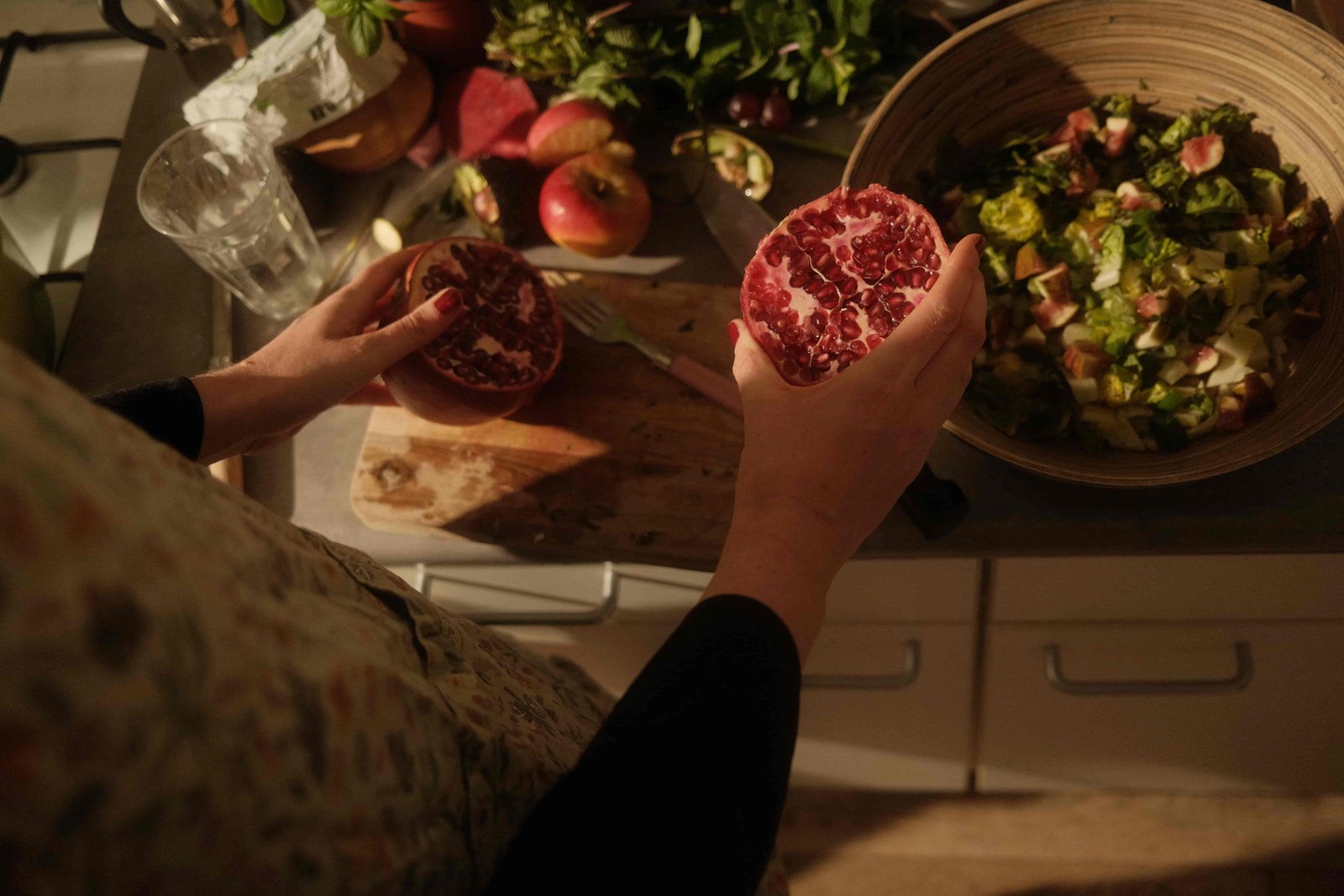 Hands holding a halved pomegranate over a wooden board with fresh herbs, fruit, and salad on a kitchen counter.